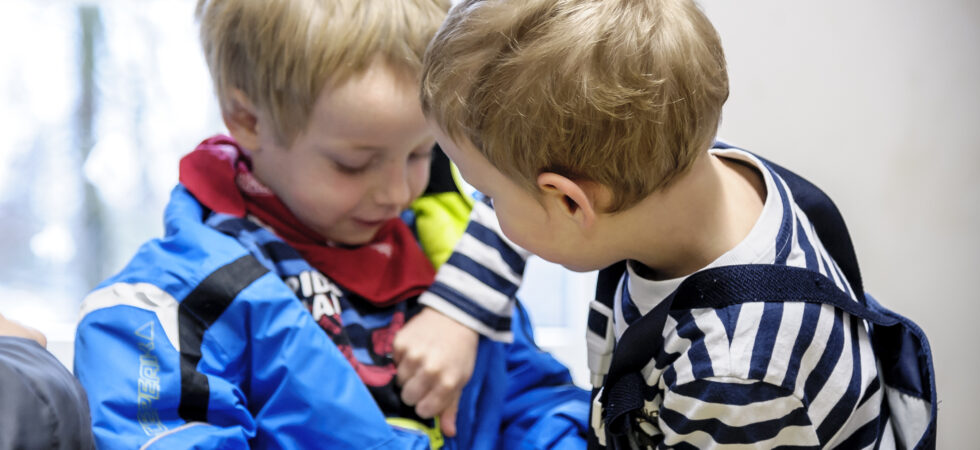 Dekoratives Bild. Zwei Kinder in Regenkleidung, einander zugewandt. Das vordere Kind hilft dem anderen Kind beim Reißvershluss schließen.